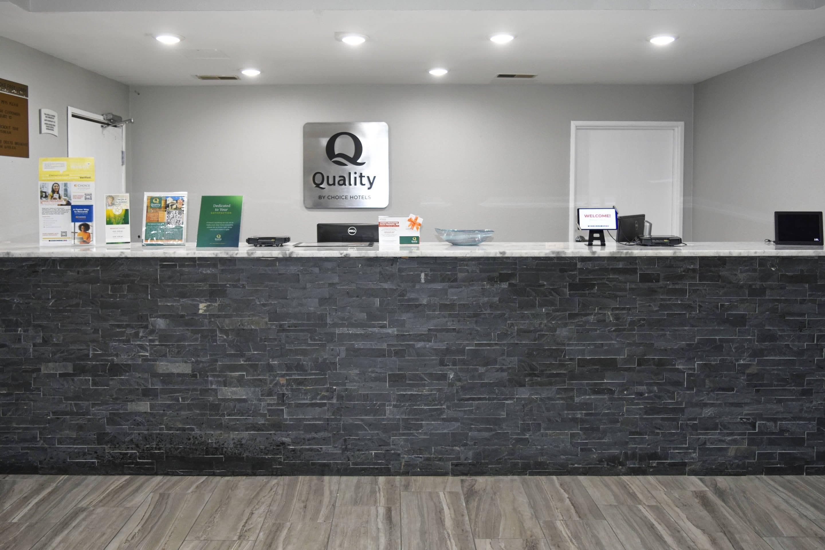 A hotel reception desk with a dark stone counter, a sign reading 'Quality by Choice Hotels', and a computer monitor displaying 'WELCOME!'.
