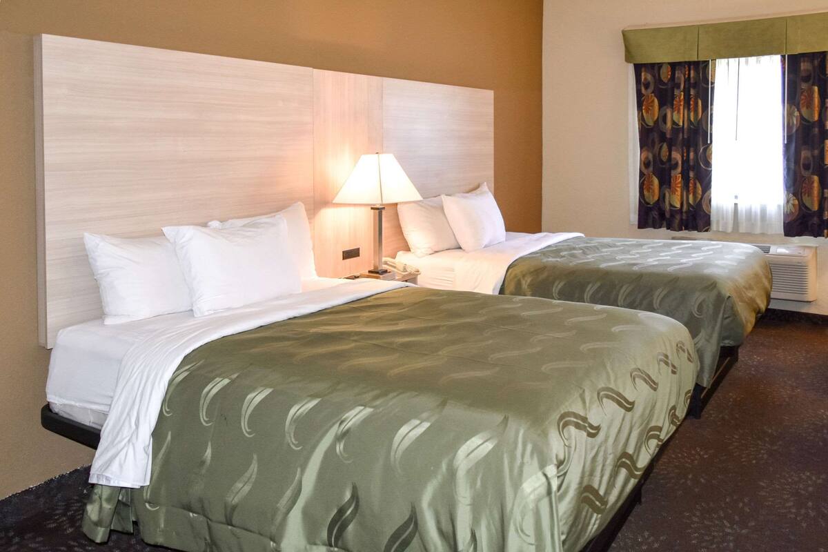 Quarto de hotel com duas camas, uma mesa de cabeceira com abajur e uma janela com cortinas.