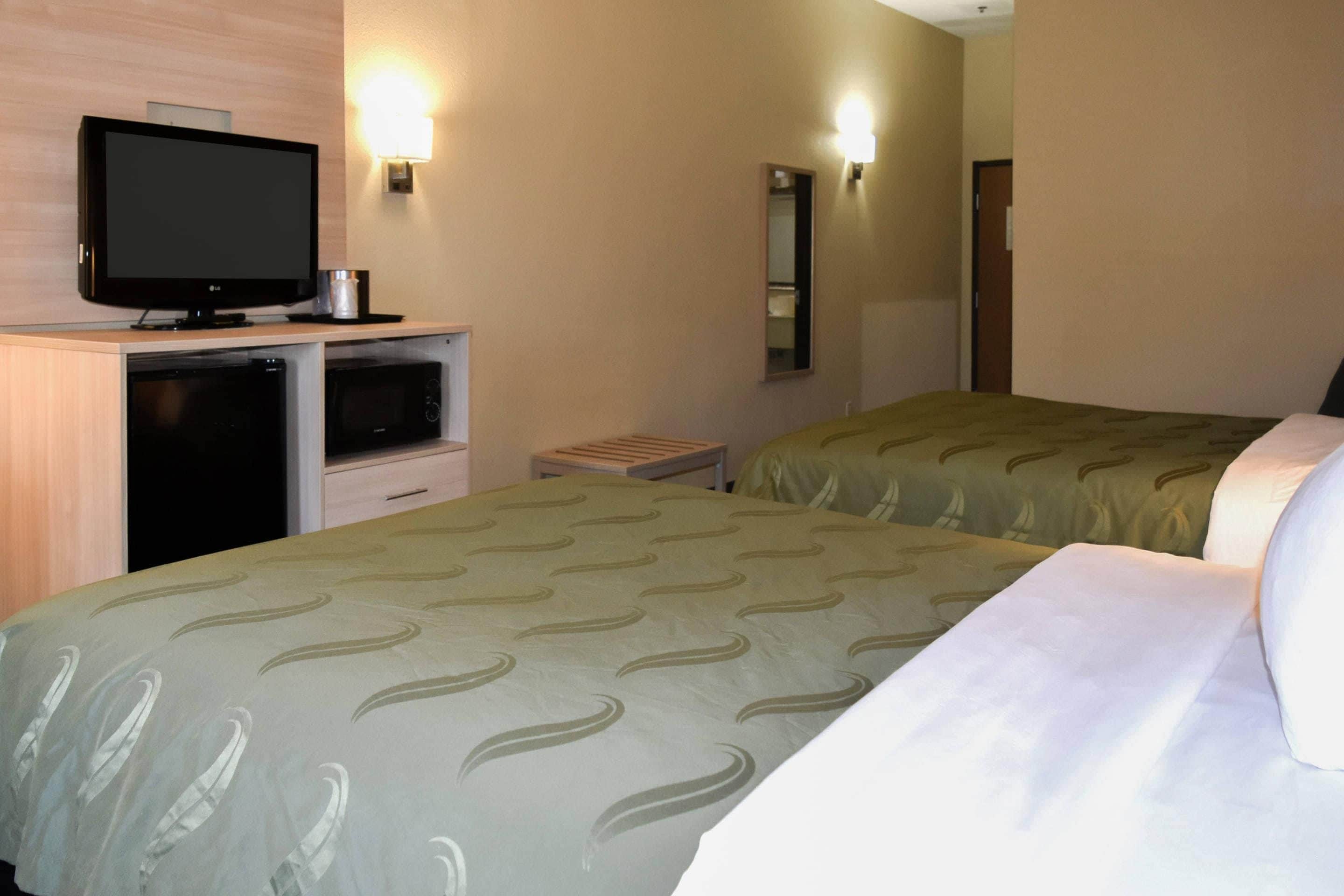 A hotel room with a television on a desk, two beds with green patterned bedding, a small table, and a mirror on the wall.