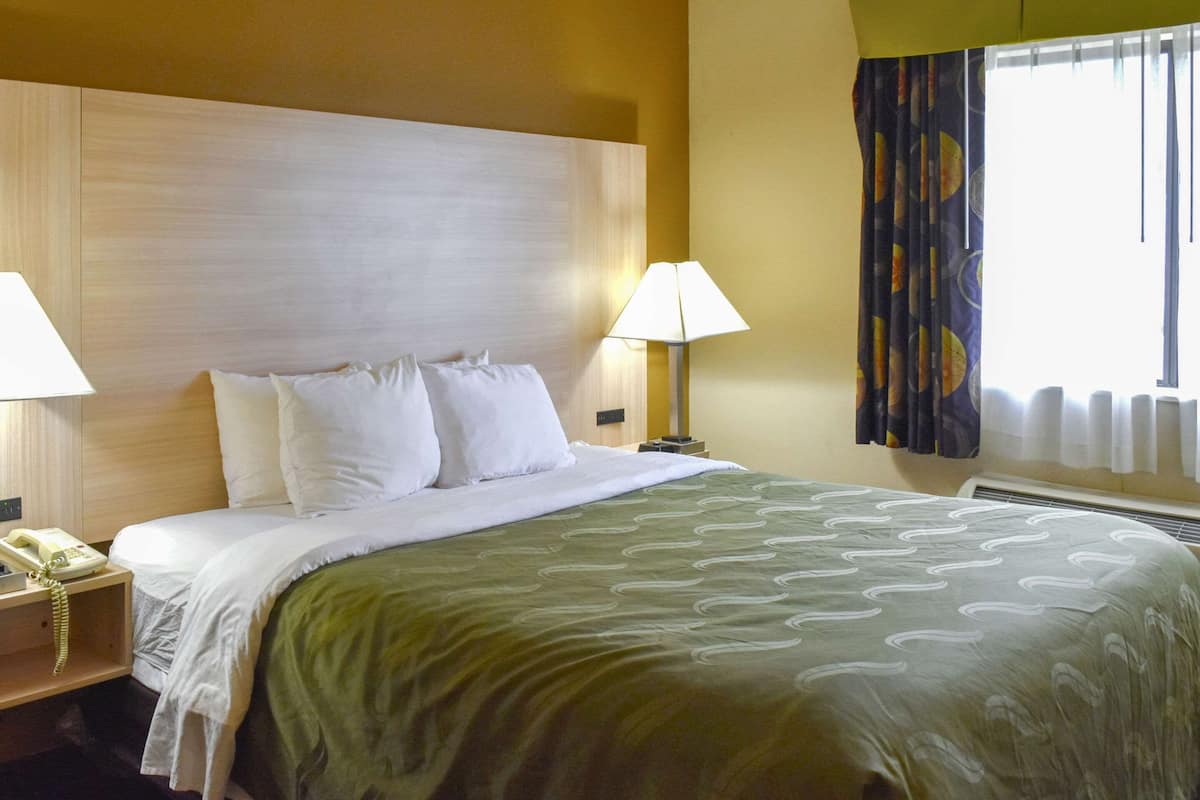 Quarto de hotel com uma cama grande, duas luminárias de cabeceira, um telefone e uma janela com cortinas.