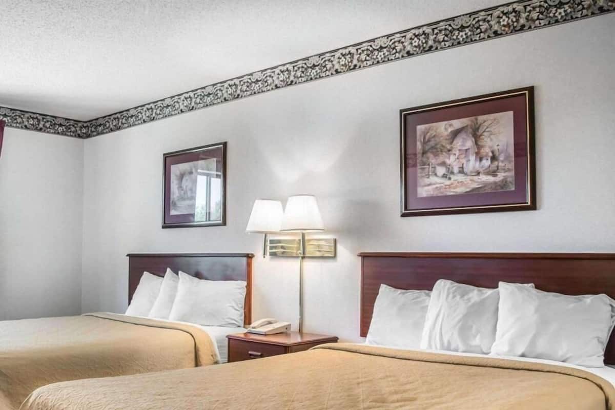 Quarto de hotel com duas camas, mesa de cabeceira com abajur e quadros nas paredes.