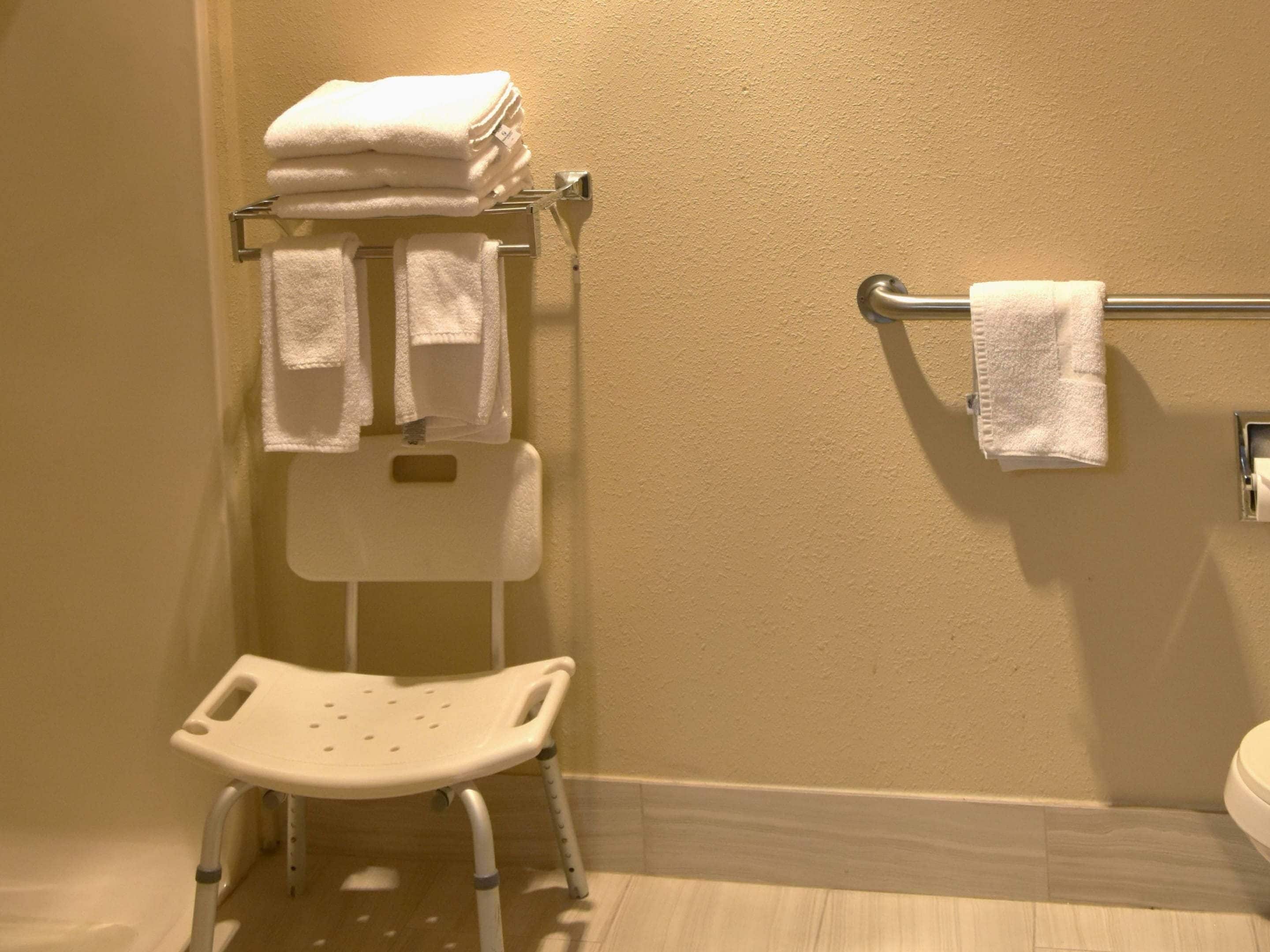A bathroom with a white toilet, a white sink, a white shower chair, and a towel rack with folded towels.