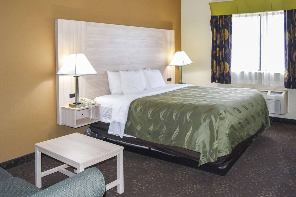Quarto de hotel com uma cama, duas mesas de cabeceira com luminárias, uma mesinha e uma janela com cortinas.