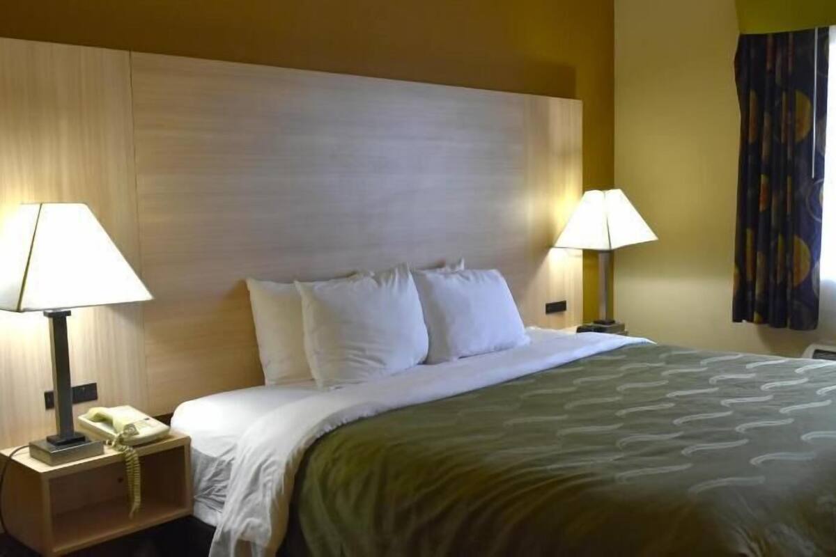 Quarto de hotel com cama, duas luminárias de cabeceira, telefone e janela com cortinas.