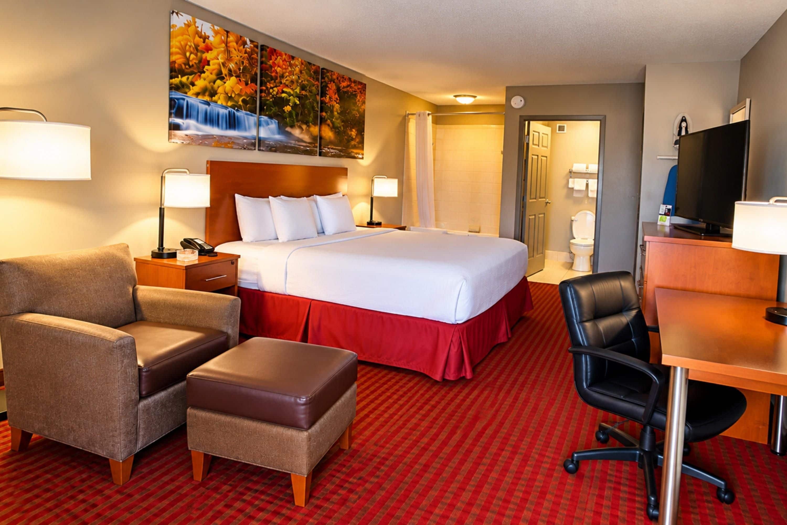 A hotel room with a bed, two bedside tables, a desk with a chair, a sofa, an ottoman, a television, and a painting on the wall.