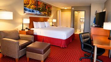 A hotel room with a bed, two bedside tables, a desk with a chair, a sofa, an ottoman, a television, and a painting on the wall.