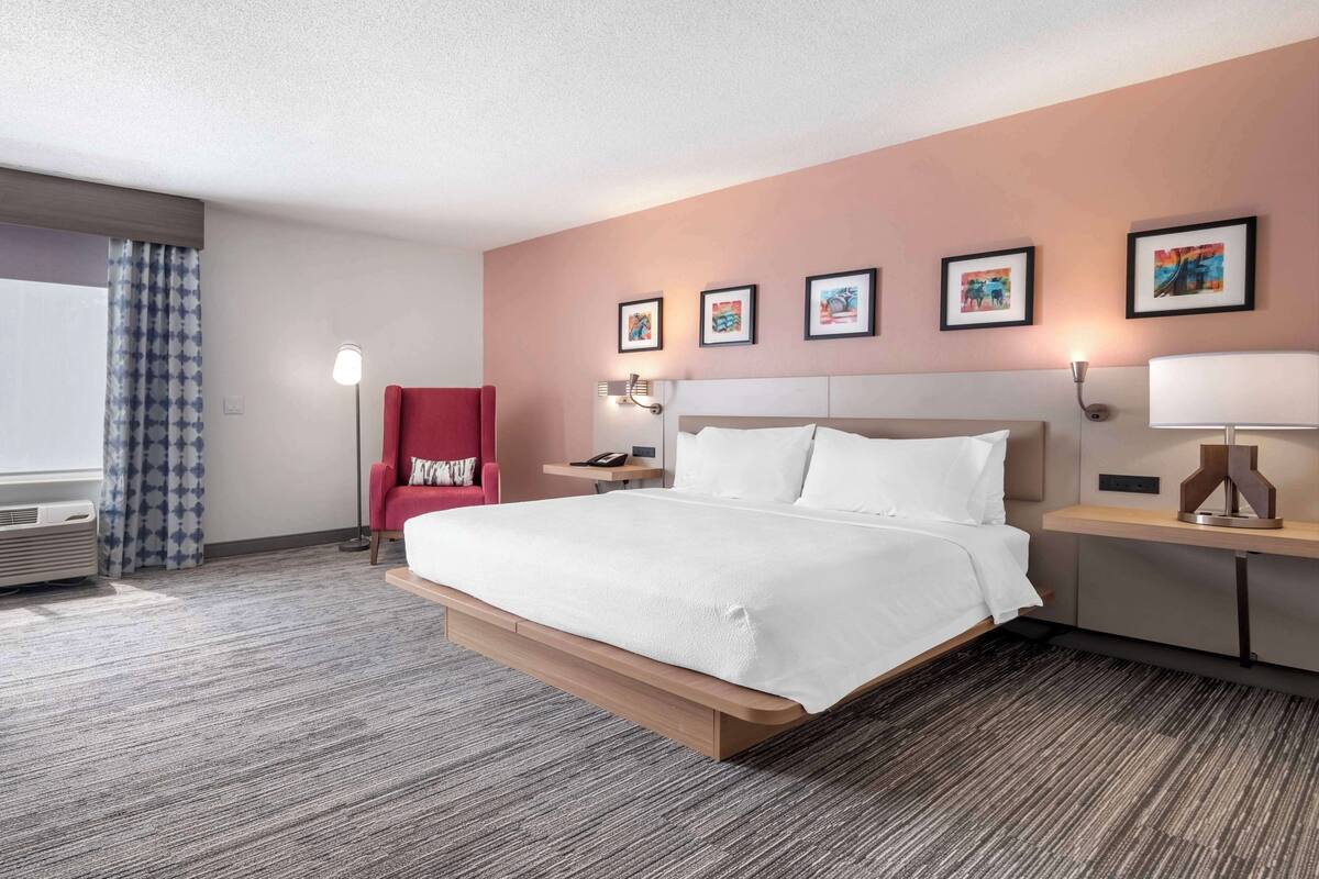 A modern hotel room with a large bed, a red armchair, a bedside table with a lamp, and framed artwork on the wall.