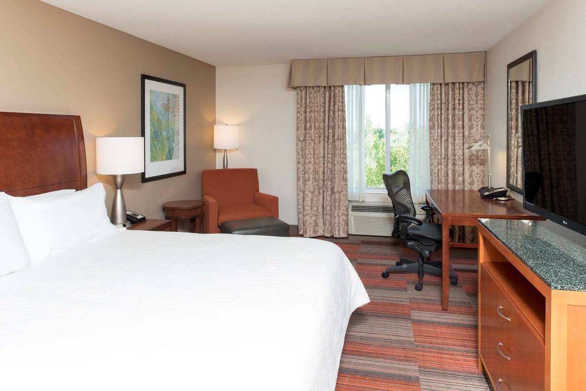 A hotel room with a bed, desk, chair, TV, lamp, and window with curtains.
