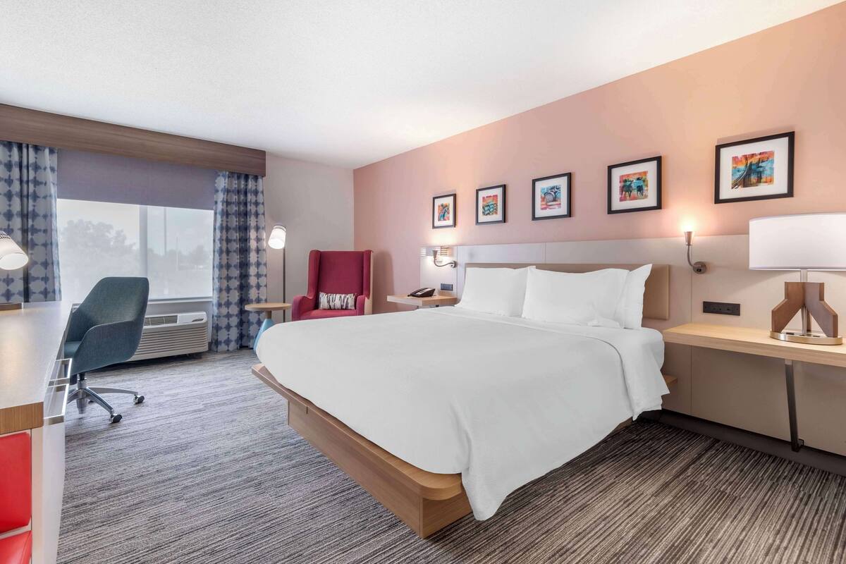 A hotel room with a large bed, a desk with a lamp, a chair, and a window with curtains.