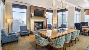A spacious room with a large wooden table, blue patterned chairs, a fireplace, and large windows offering views of the outdoors. - Hilton Garden Inn Lexington Hamburg (Lexington)
