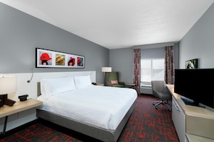 Habitación de hotel con cama, escritorio, silla, televisor y ventana con cortinas.