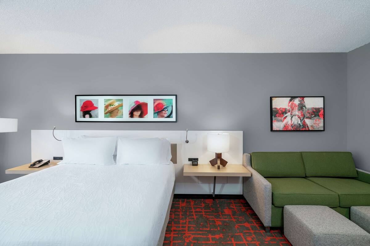 A hotel room with a bed, a green sofa, a nightstand with a lamp, and two framed pictures on the wall.