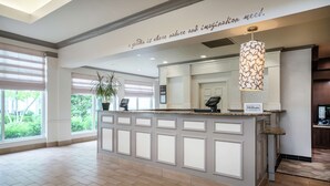 Zona de recepción de hotel con un mostrador grande, una planta en maceta y un cartel que indica la marca Hilton.