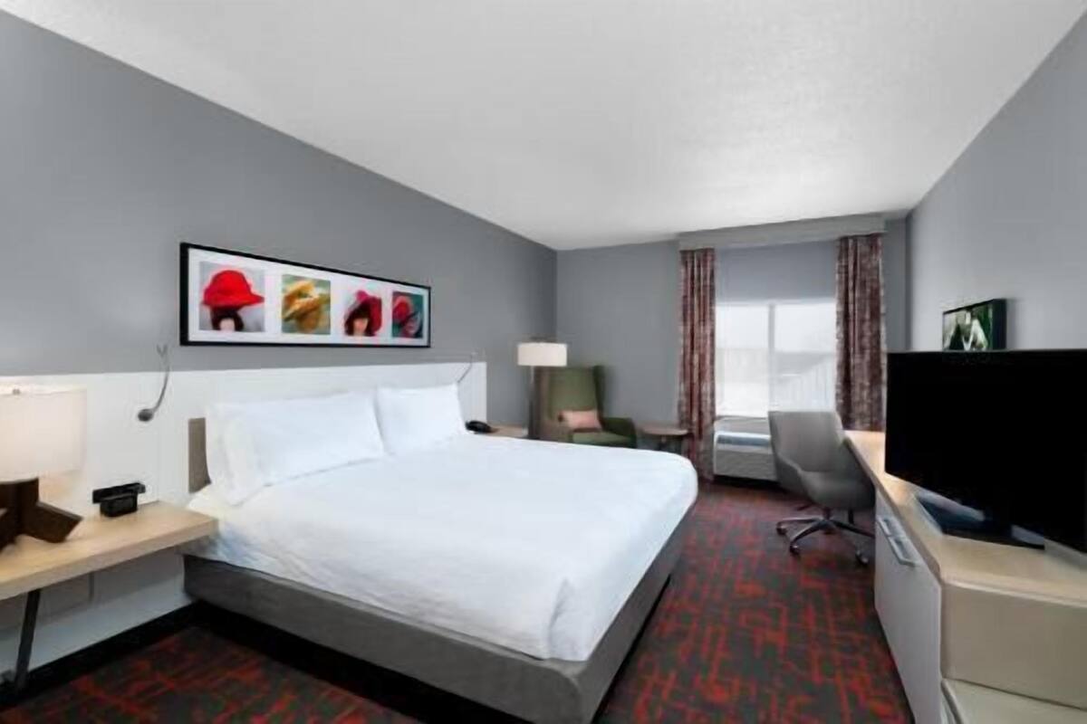 A hotel room with a bed, a desk, a chair, a TV, and a framed picture on the wall.