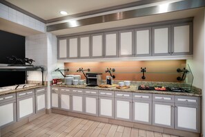 Une cuisine moderne avec des meubles gris, un four encastré et un îlot central doté d’une plaque de cuisson en verre et de divers ustensiles de cuisine.