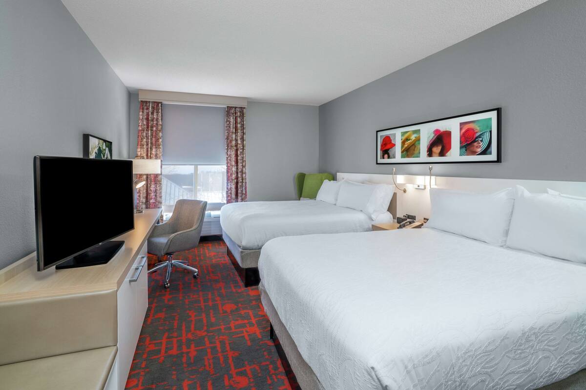 A hotel room with two beds, a TV, a desk, and a window with curtains.