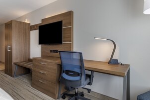 Una habitación de hotel moderna con un escritorio de madera, un televisor de pantalla plana, una silla de oficina de malla azul, una lámpara de escritorio y un armario.