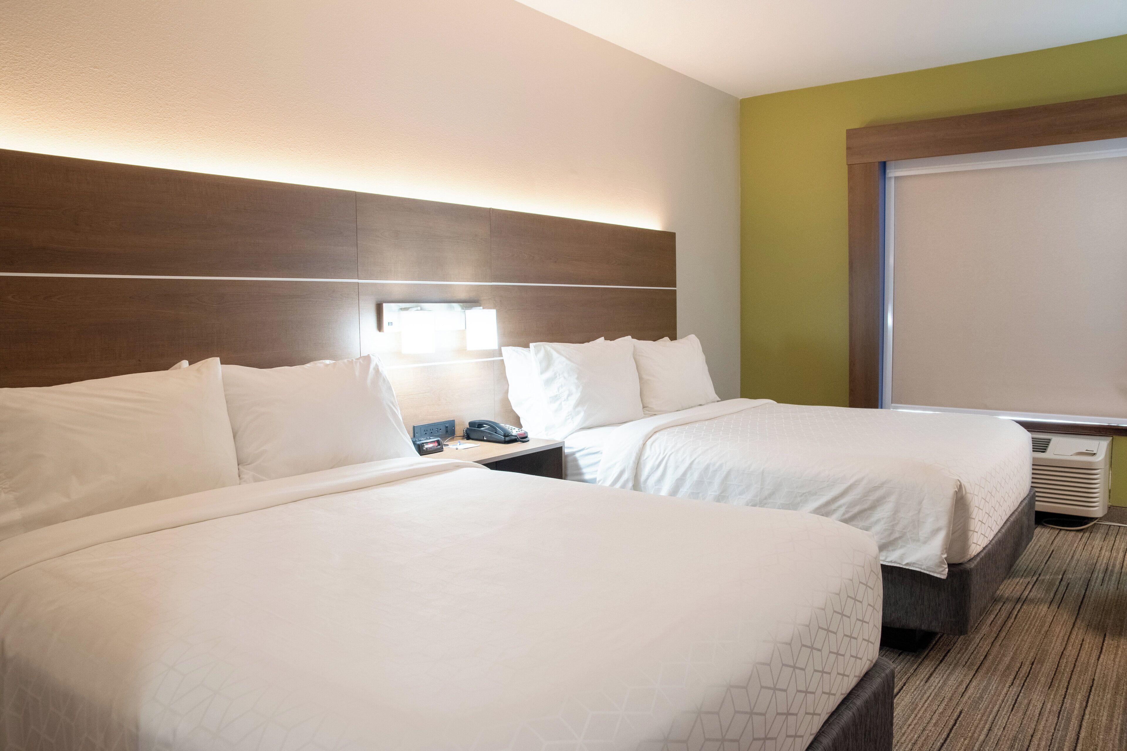 A hotel room with two beds, a bedside table, a lamp, and a window.