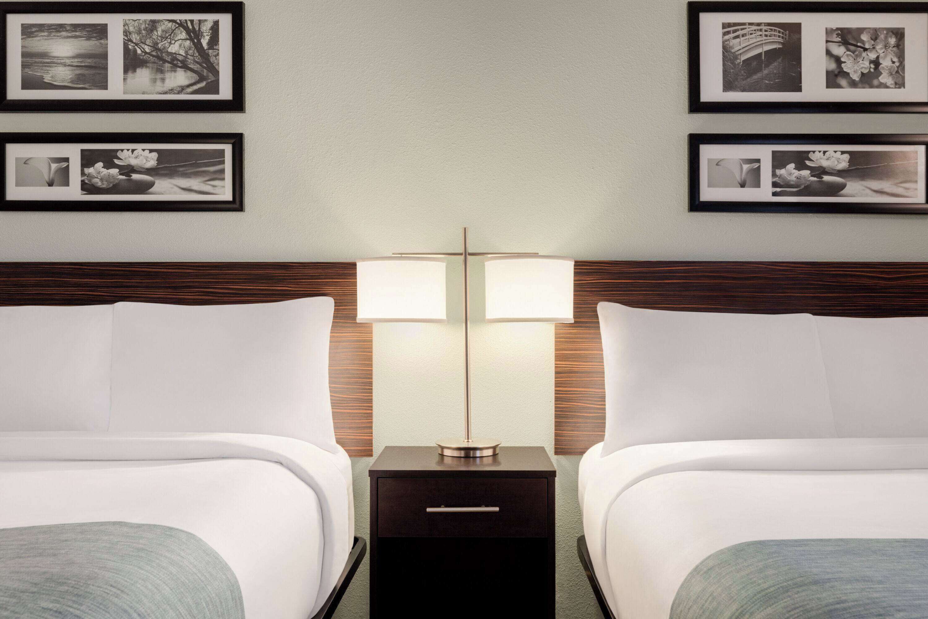 A hotel room with two beds, a nightstand with two lamps, and framed pictures on the wall.