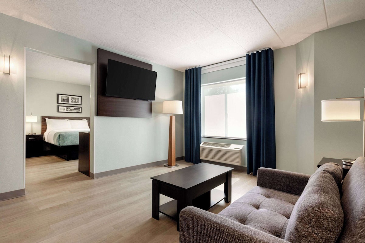 Habitación de hotel con cama, televisión montada en la pared, escritorio con lámpara, sofá y ventana con cortinas.