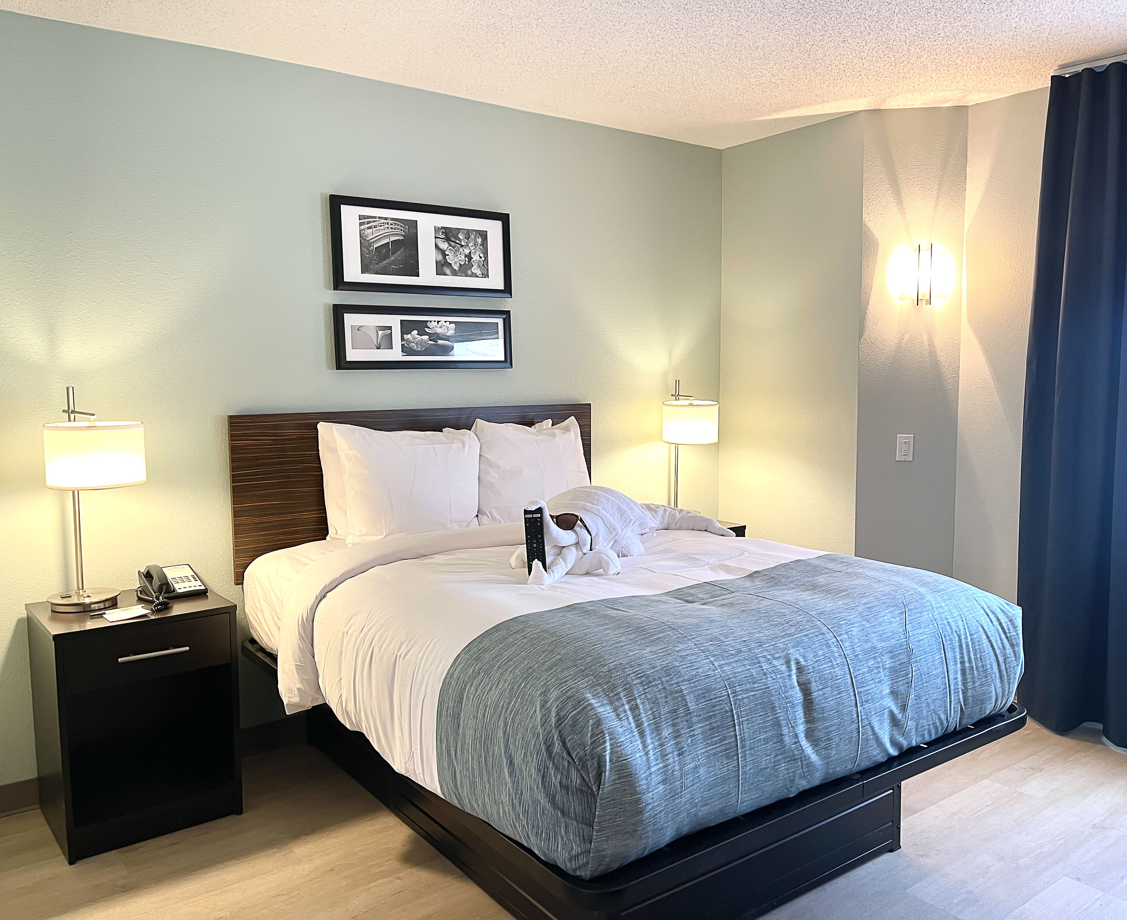 A neatly made bed with white and gray bedding, two bedside lamps, a nightstand with a phone, and a framed picture above the bed.