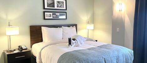 A neatly made bed with white and gray bedding, two bedside lamps, a nightstand with a phone, and a framed picture above the bed.