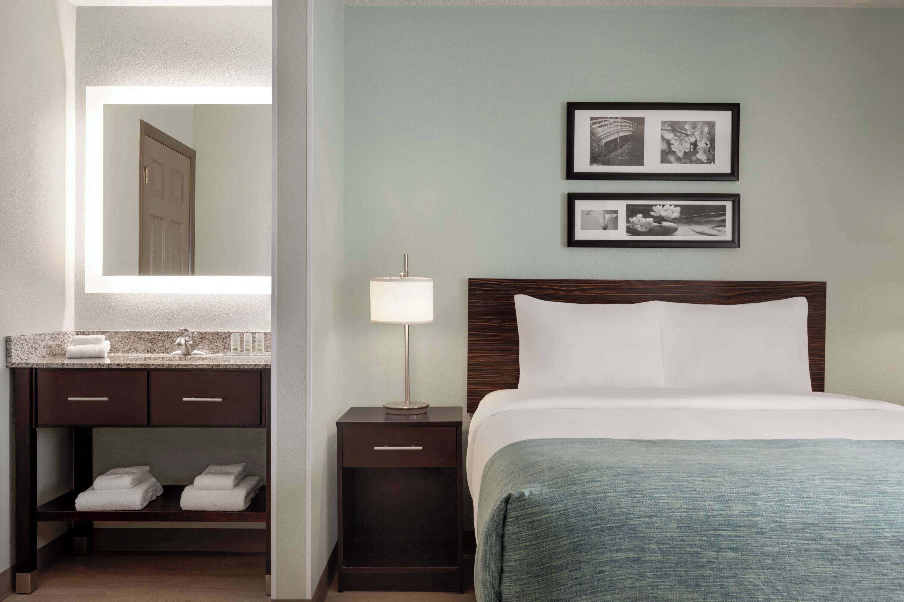 A hotel room with a bed, a nightstand with a lamp, a vanity with a mirror, and two framed pictures on the wall.