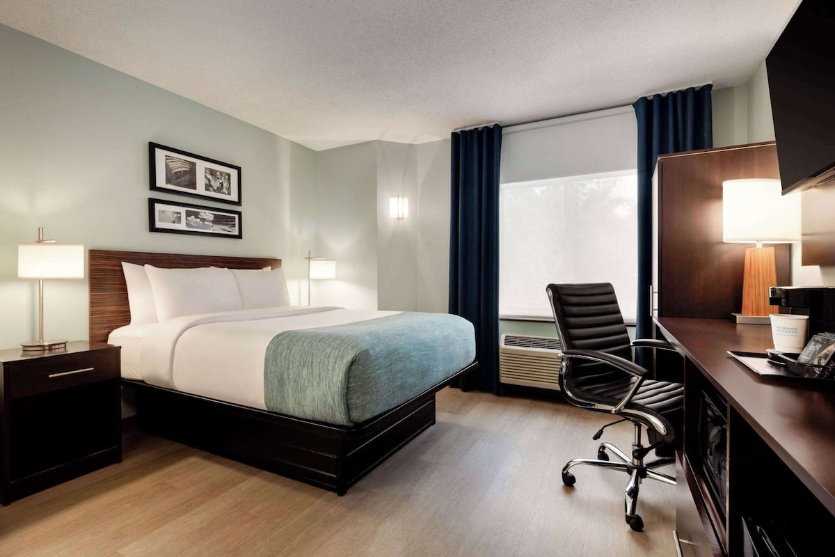 Habitación de hotel con cama, mesitas de noche, escritorio con silla, ventana con cortinas e iluminación en la pared.
