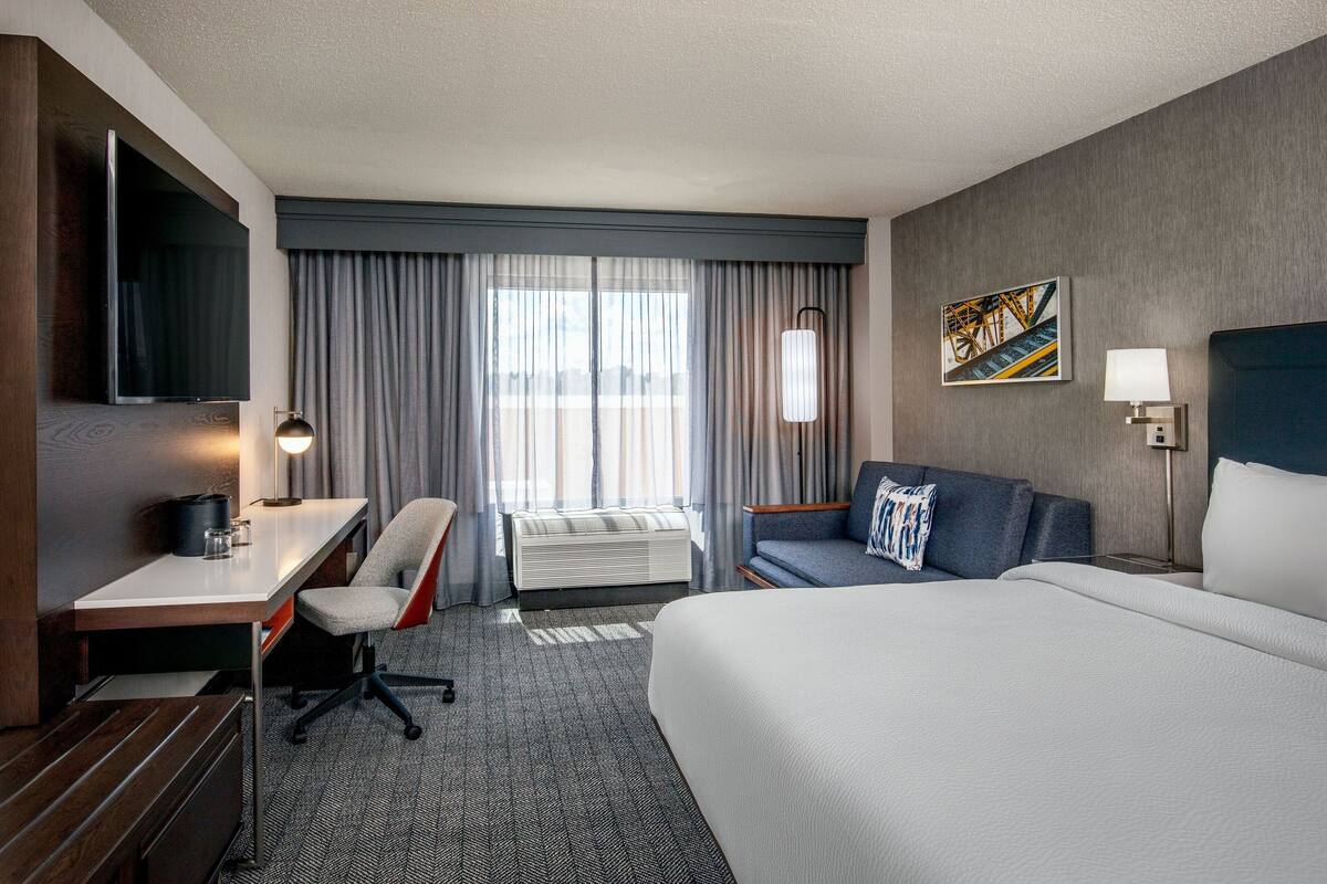 A hotel room with a large bed, a desk with a chair, a flat-screen TV, a window with curtains, and a painting on the wall.