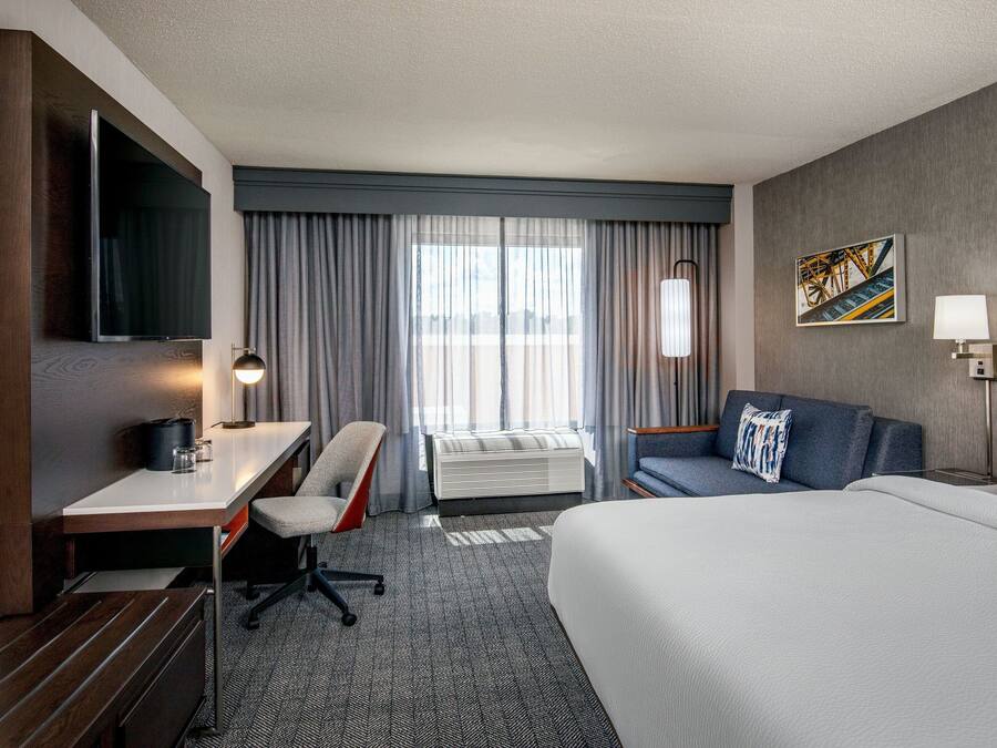 A hotel room with a large bed, a desk with a chair, a flat-screen TV, a window with curtains, and a painting on the wall.