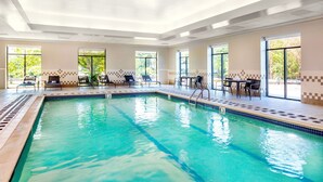 An indoor swimming pool with clear blue water, surrounded by lounge chairs and tables, large windows with a view of greenery, and a tiled floor.