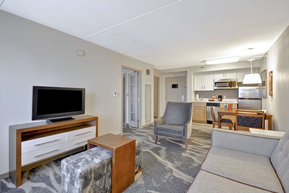 A modern hotel room with a flat-screen TV, a sofa, a small table, and a kitchenette.