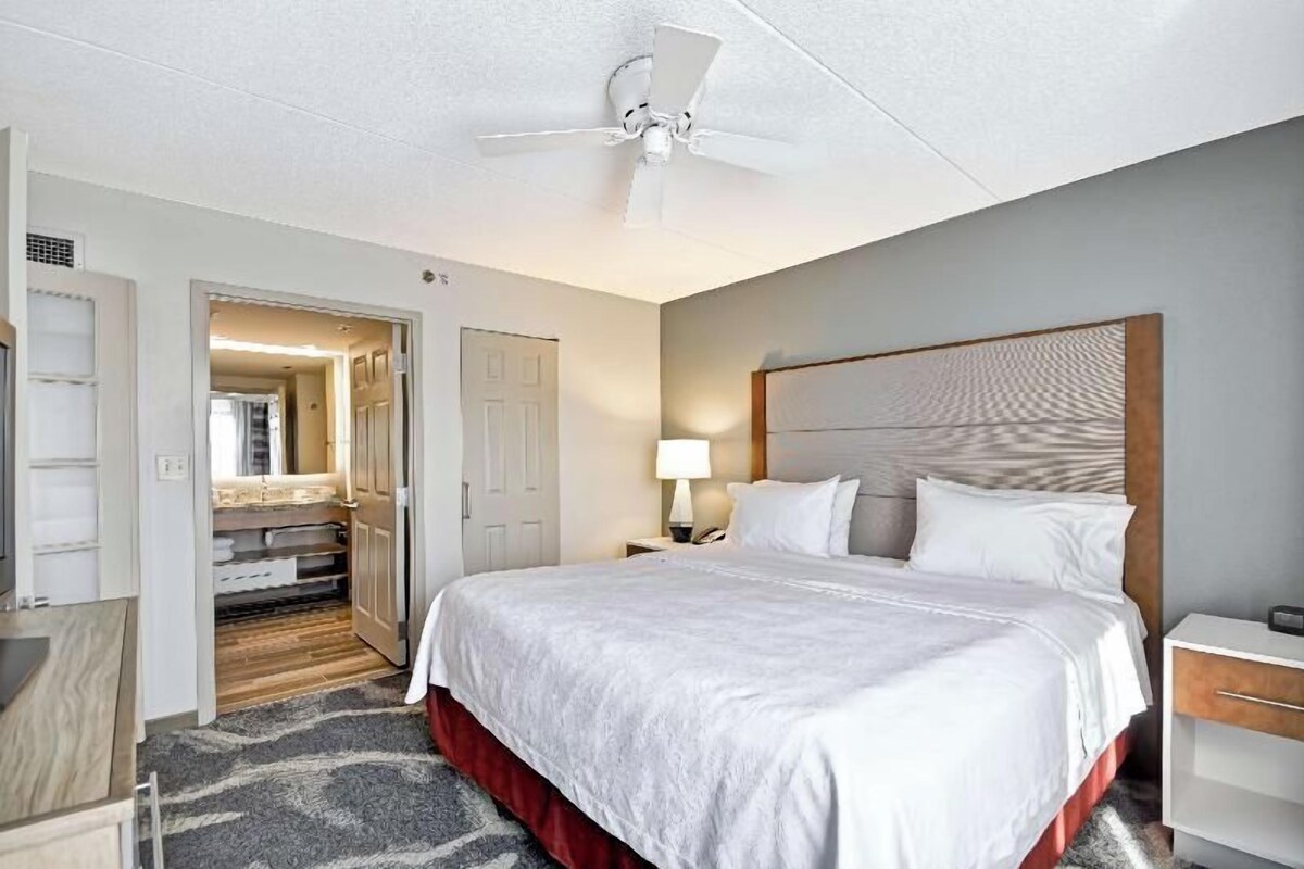 A hotel room with a bed, a nightstand, a lamp, a ceiling fan, and a door leading to a bathroom.