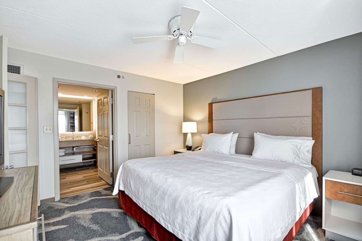 A hotel room with a bed, a nightstand, a lamp, a ceiling fan, and a door leading to a bathroom.
