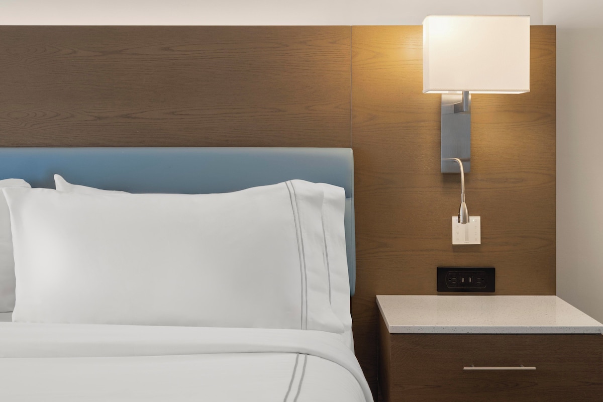A hotel room with a bed, a nightstand, a wall-mounted lamp, and a power outlet.