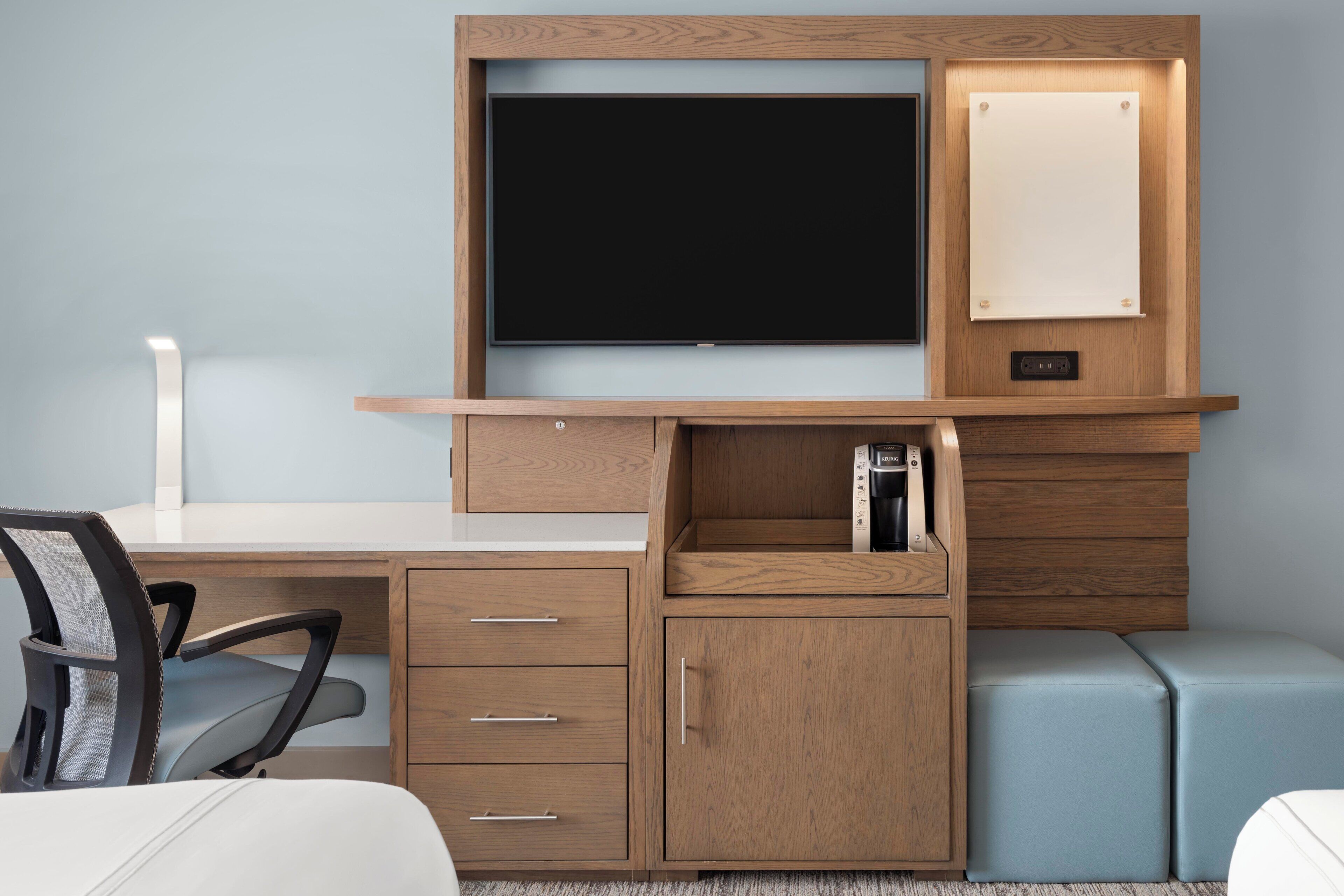 A hotel room with a wooden desk, a television, a coffee machine, a chair, and a small seating area.