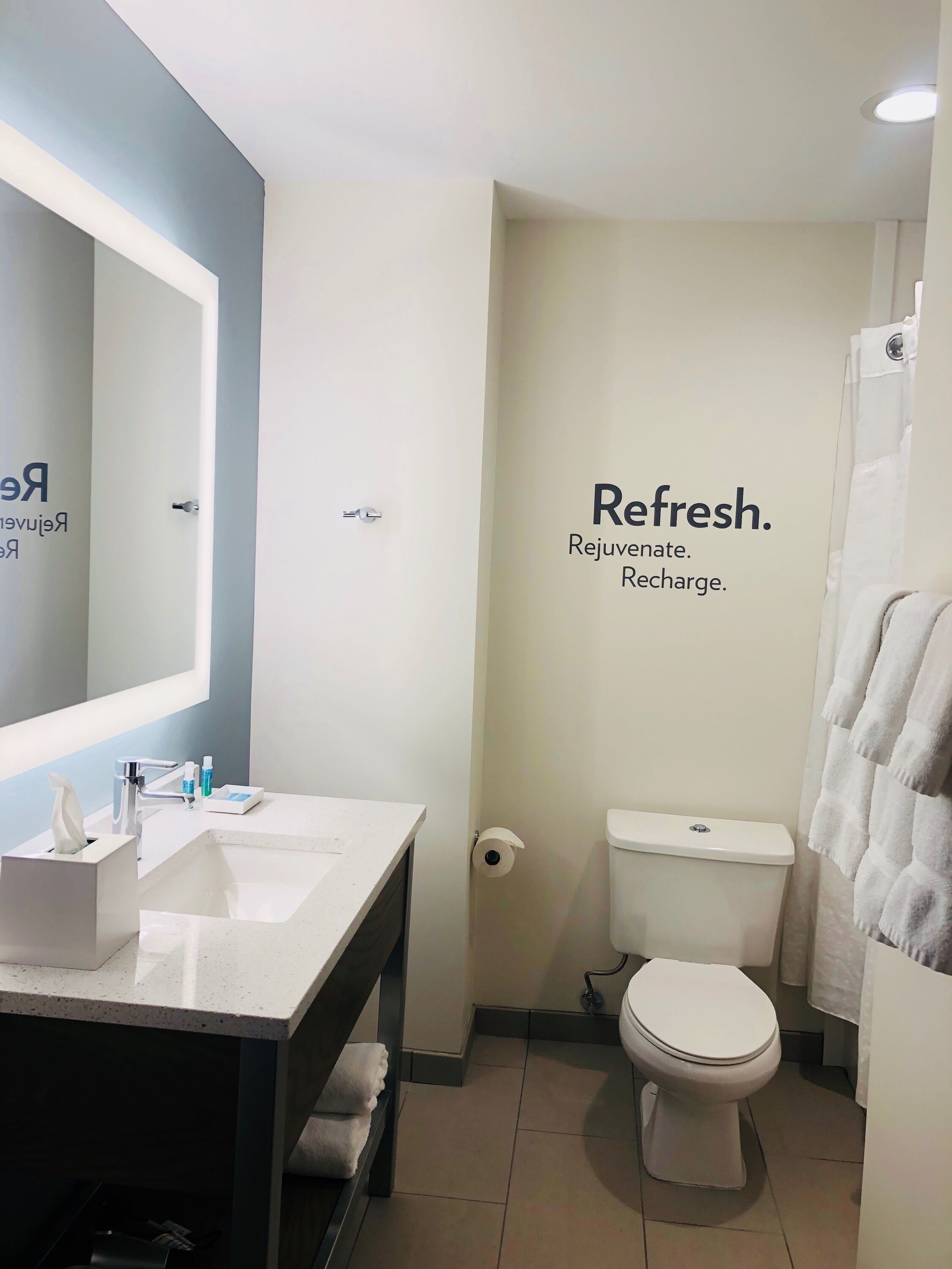 Un baño moderno con lavamanos, espejo y retrete blancos. Se ve un perchero, y las paredes tienen un diseño minimalista con un cartel que dice 'Refresh'.