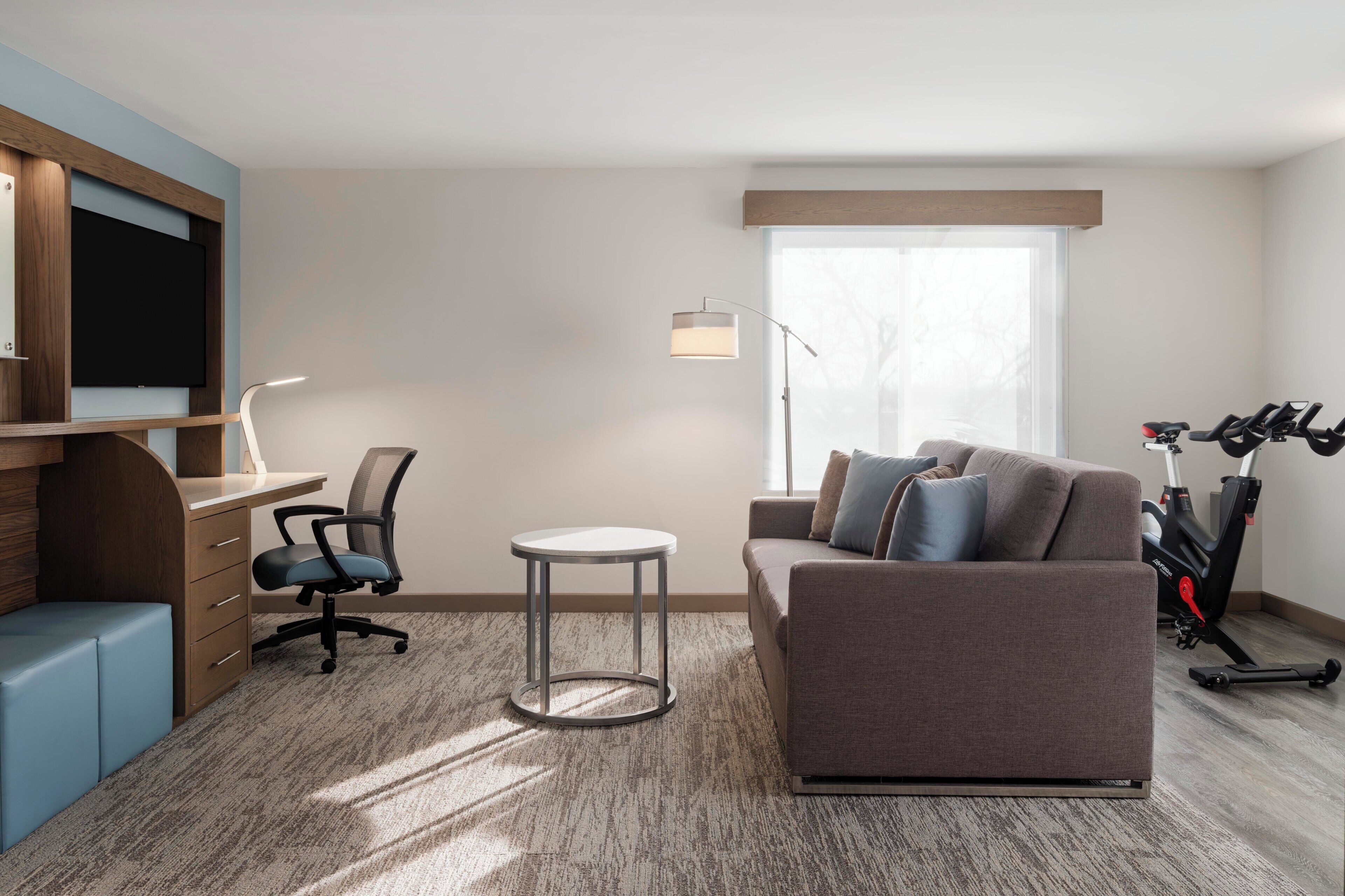 Moderni hotellihuone, jossa on työpöytä, tuoli, sohva ja kuntopyörä.