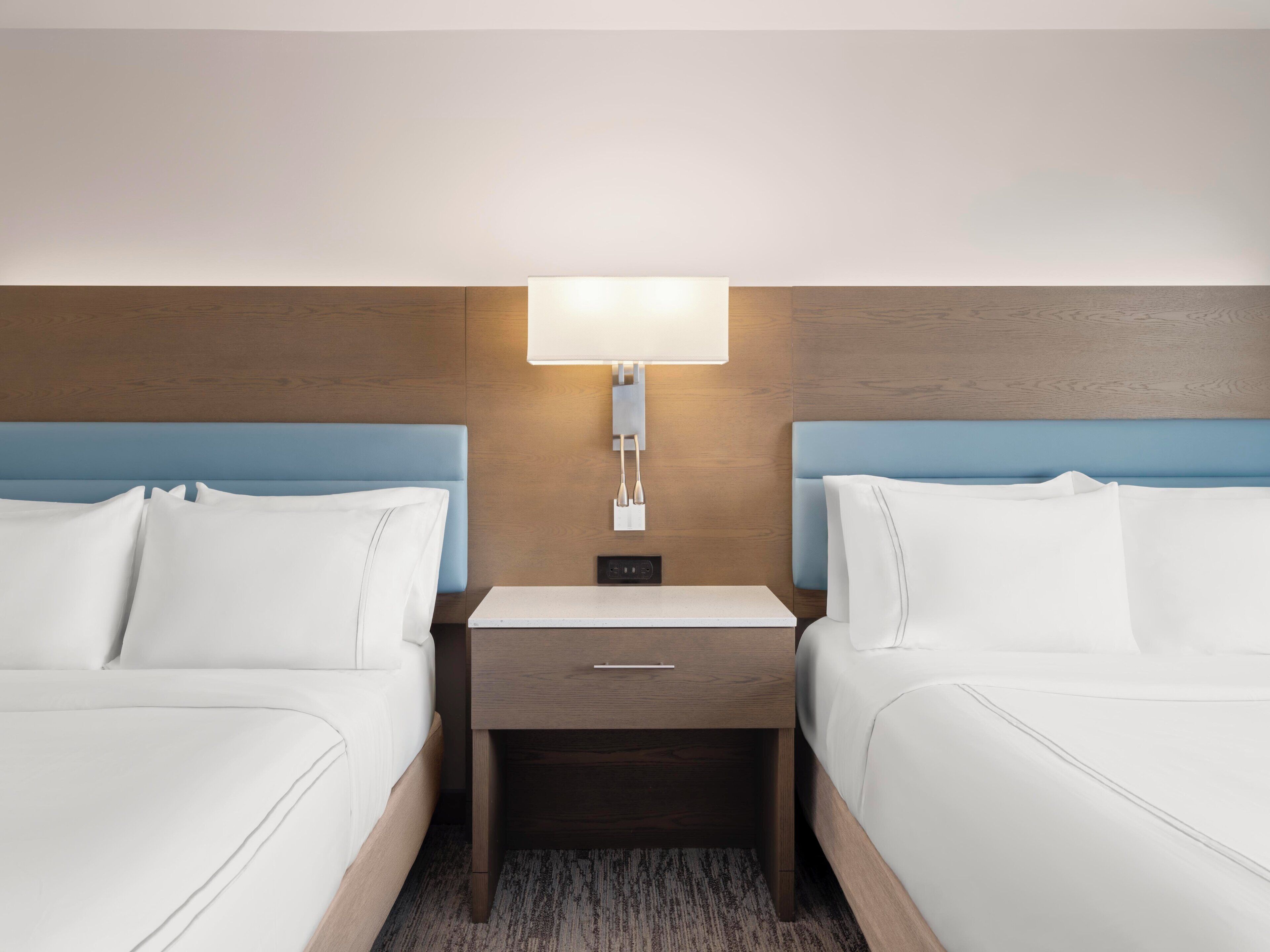 Una habitación de hotel con dos camas, una mesita de noche con lámpara y un cajón pequeño.