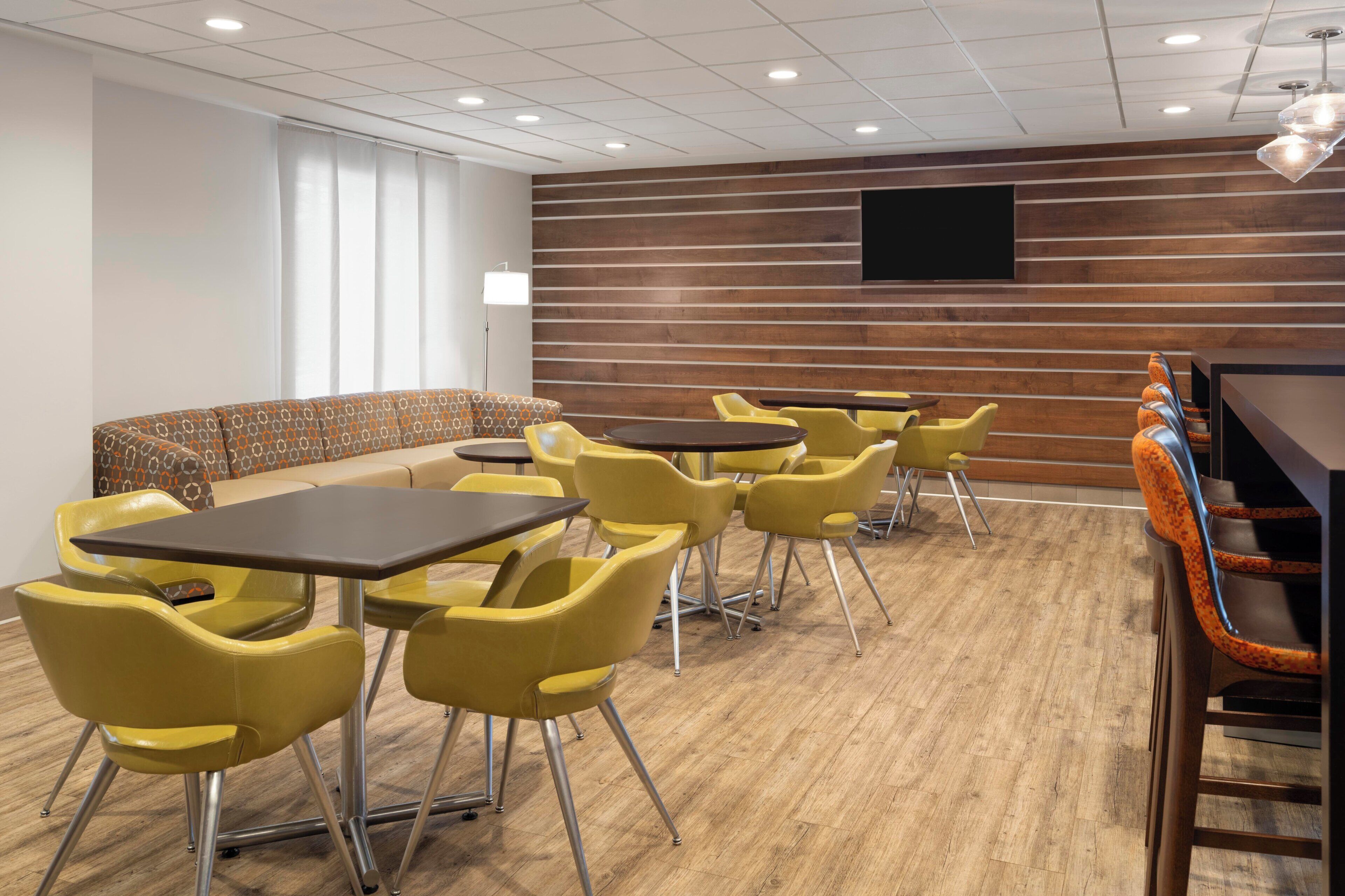 Une salle de conférence avec des chaises jaunes, un canapé marron et un mur en bois sur lequel est accroché un téléviseur à écran plat.