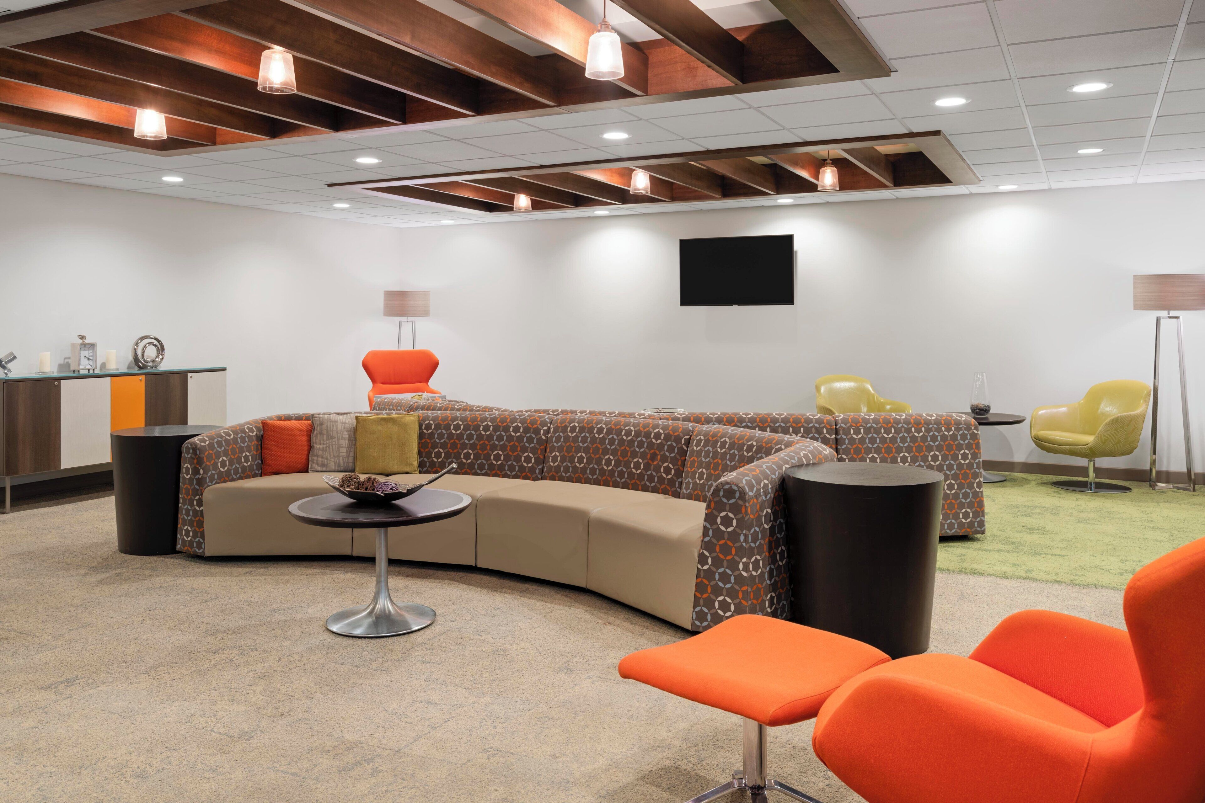A modern lounge area with a curved sofa, orange chairs, a small round table, and a flat-screen TV mounted on the wall.