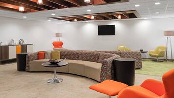 A modern lounge area with a curved sofa, orange chairs, a small round table, and a flat-screen TV mounted on the wall.