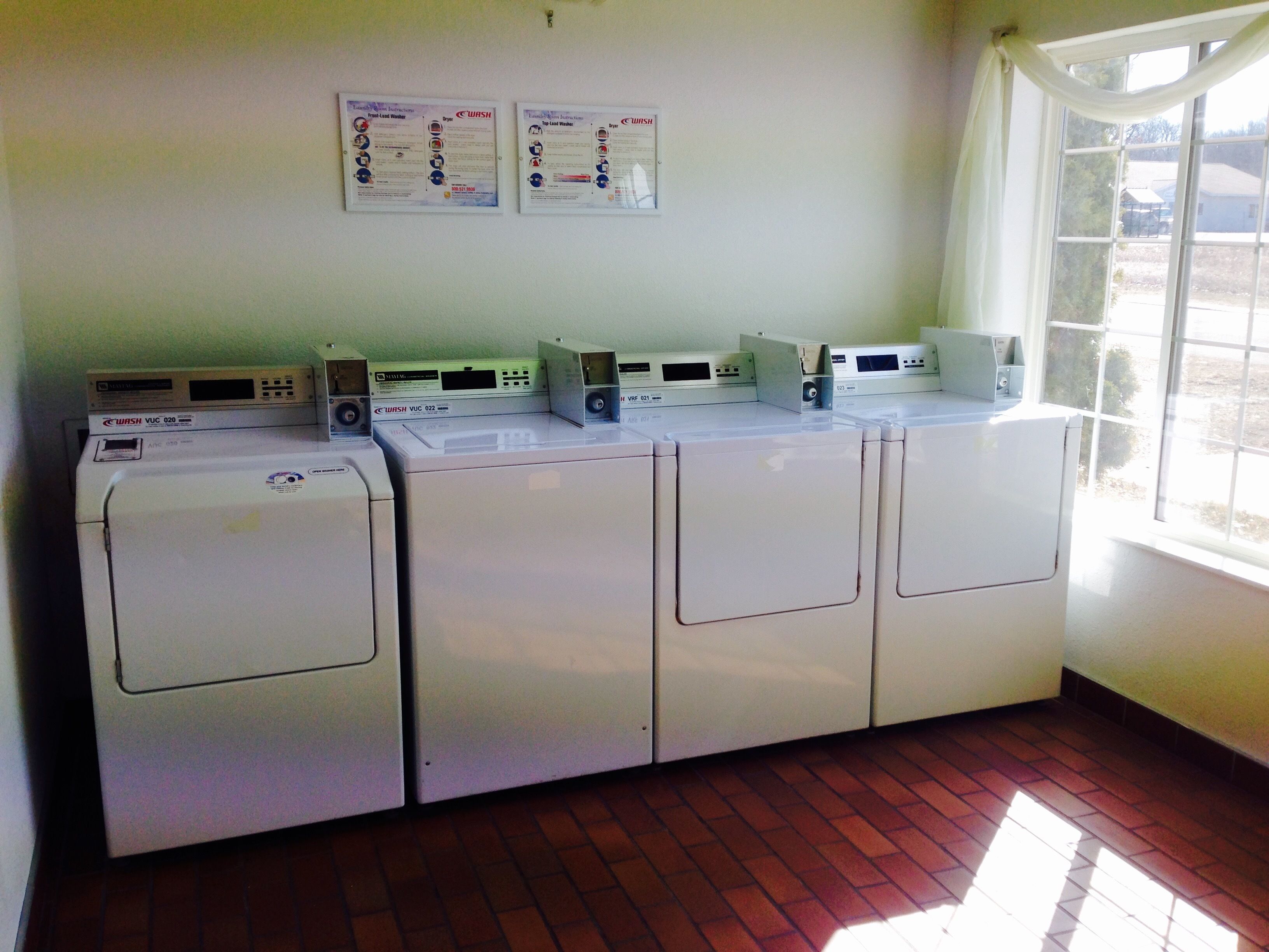 A laundry area with four washing machines and two dryers, a window with curtains, and two informational posters on the wall.