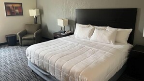 A hotel room with a large bed, a chair, a nightstand with a lamp, and a trash bin. - American Inn and Suites Houghton Lake (Houghton Lake)
