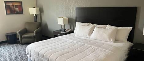 A hotel room with a large bed, a chair, a nightstand with a lamp, and a trash bin.