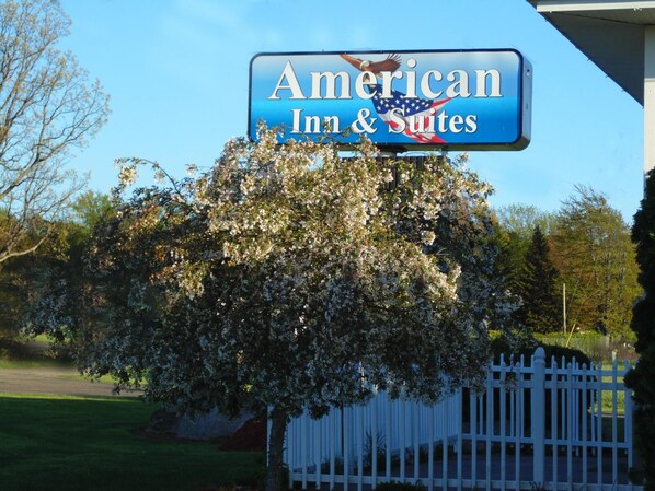 A sign for American Inn & Suites with a clear view of the surrounding area including a white picket fence and blooming trees.