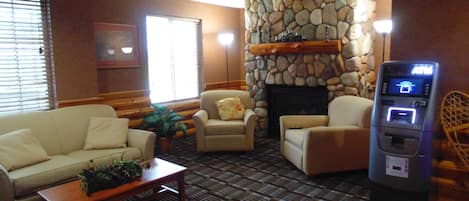 A hotel lobby with a stone fireplace, a seating area with a sofa and armchairs, a small table with a plant, and an ATM machine.