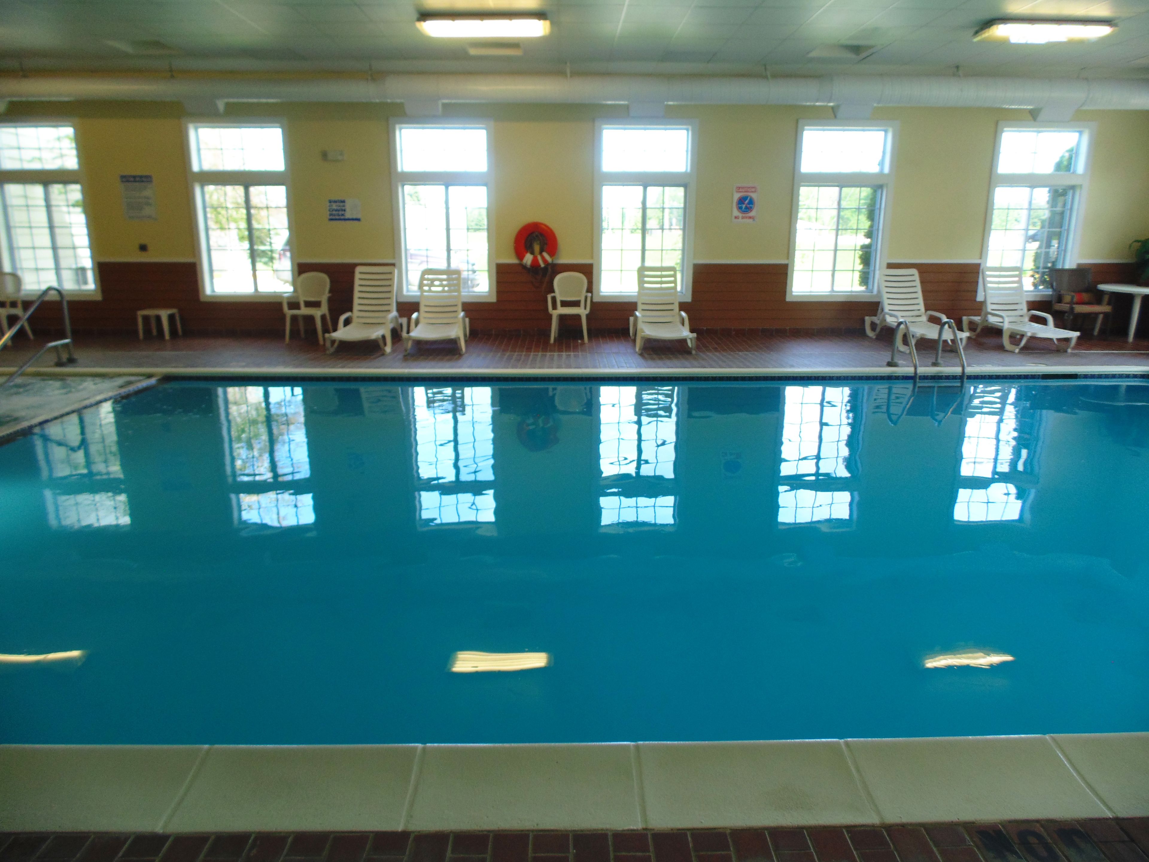 An indoor swimming pool with clear blue water, surrounded by white lounge chairs and a tiled floor.