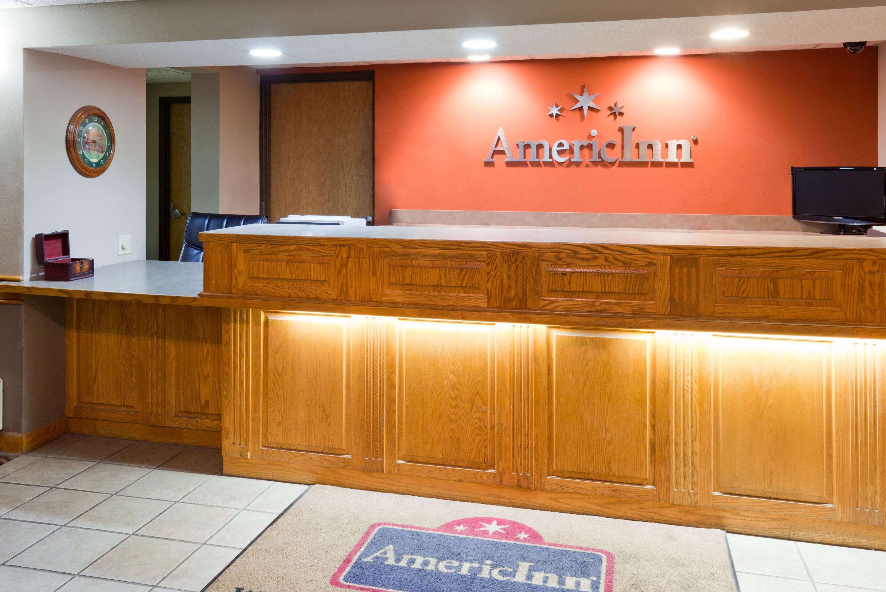 A reception desk with a wooden finish, a clock on the wall, and a carpet with the American Inn logo.
