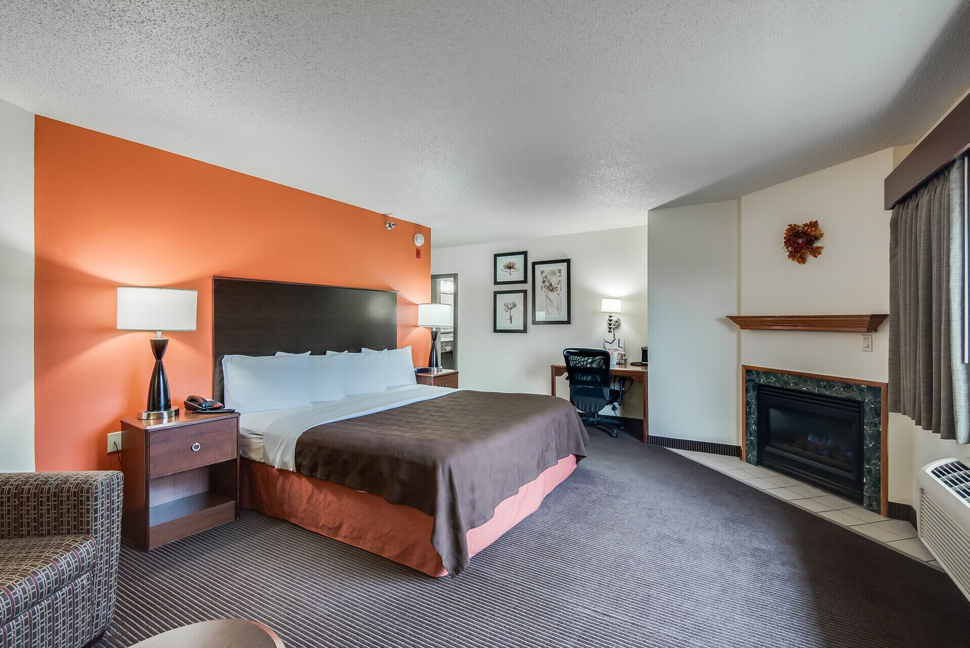 A hotel room with a bed, bedside tables, a desk, a chair, a fireplace, and framed artwork on the wall.